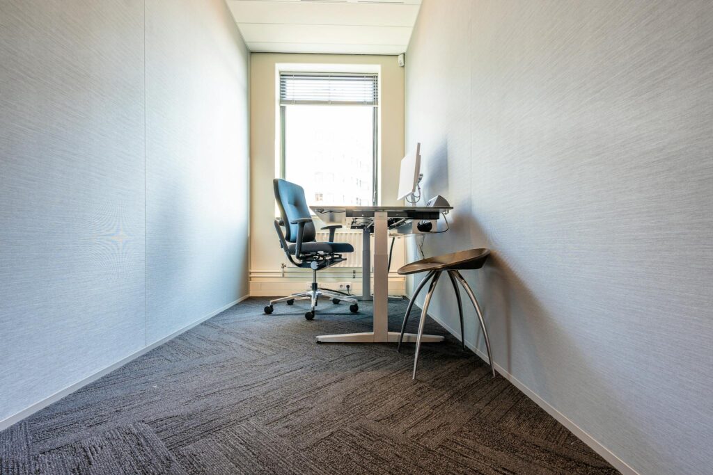 A clean, minimalist office space featuring a desk, chair, and computer by a large window.